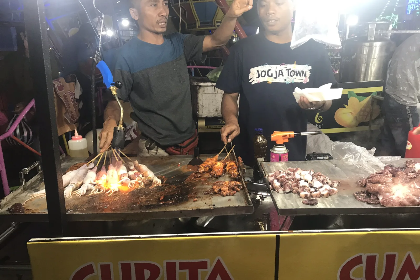 Cooks grilling skewers under the lamps of a stall at the Ubud night market
